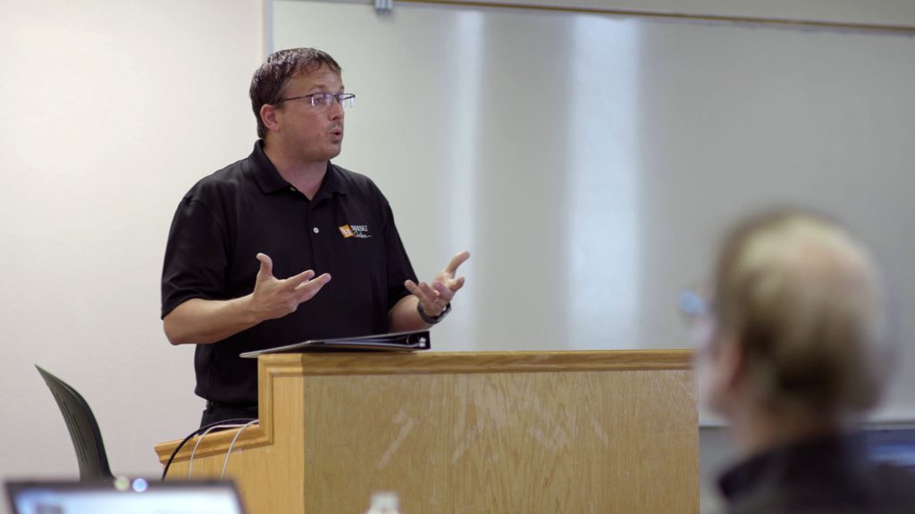 A man wearing glasses and a black SHSU Online polo shirt stands behind a wooden podium, gesturing with both hands as he speaks to an audience. A large white screen or board is behind him, and the blurry back of an audience member's head is visible in the foreground